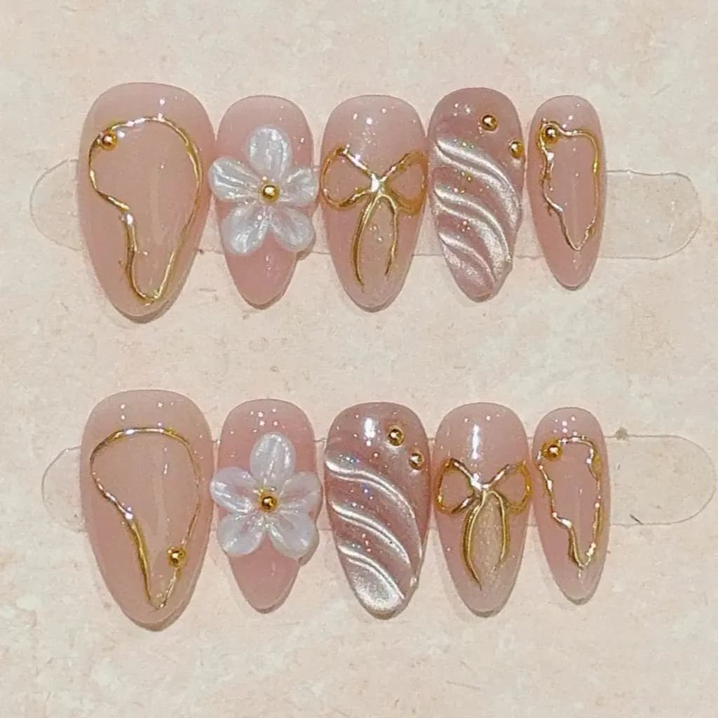 Set of 10 nude pink short almond press-on nails with 3D white flowers and gold metallic bow designs.