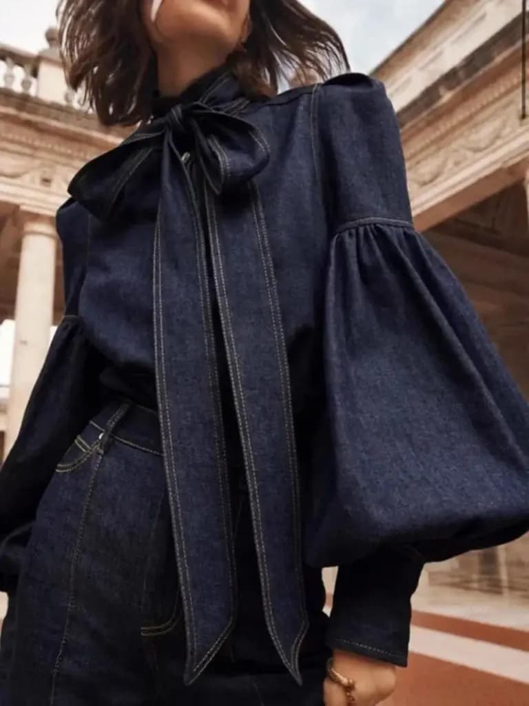 Woman wearing a dark wash imitation denim blouse with large puff sleeves and a bow-tied high neck.